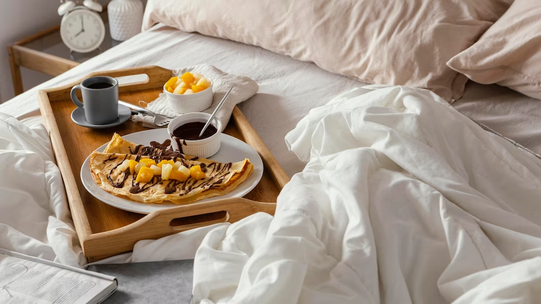fruits, a cup of drink, and a crepe arranged on a tray resting on a bed.